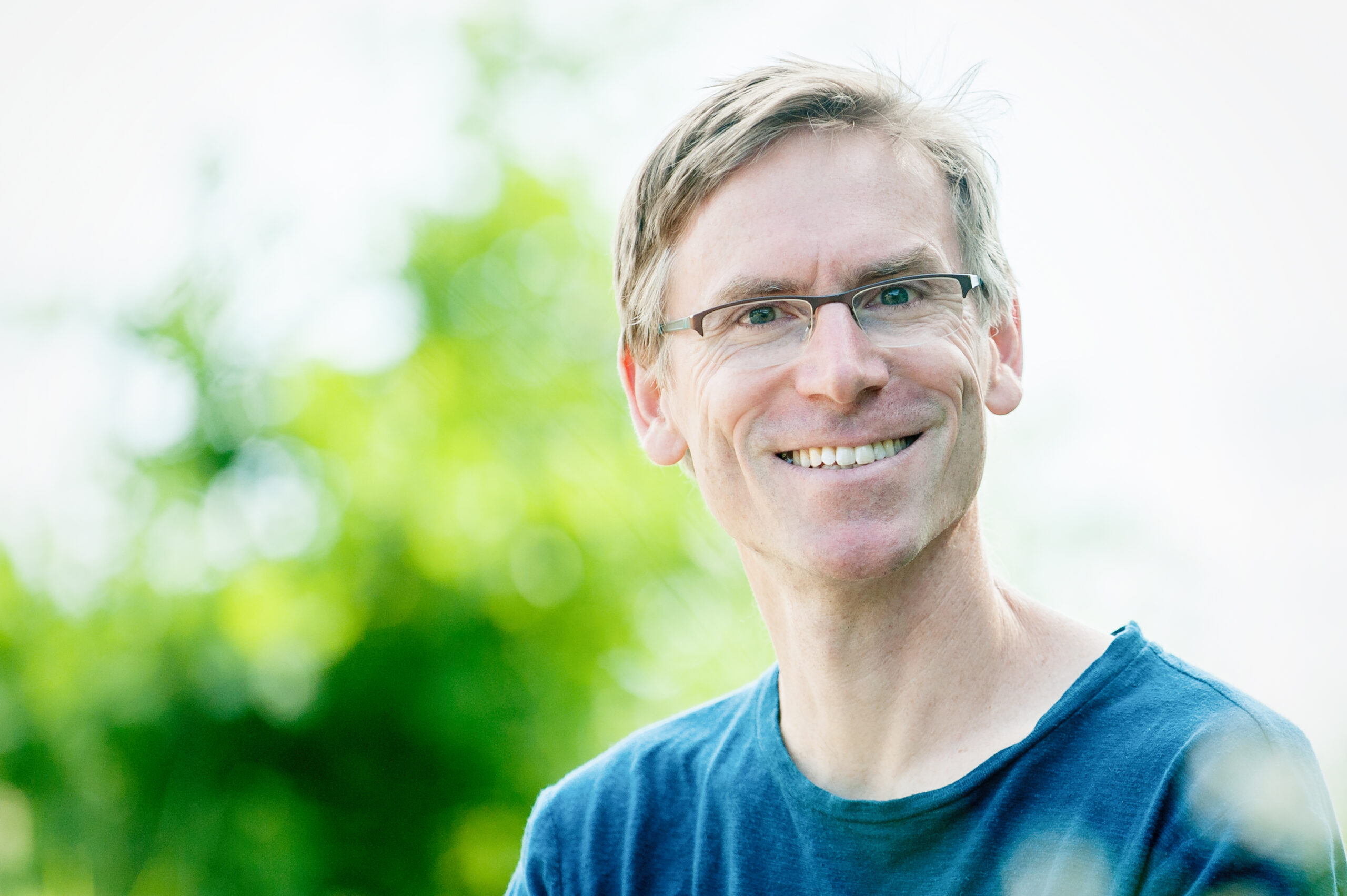 Lächelnder Mann mit Brille, grauen Haaren und blauem Pullover vor grün weißem Hintergrund