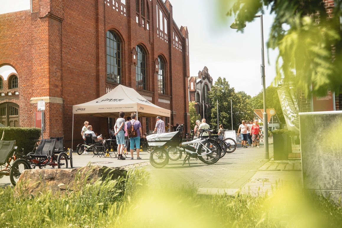 Mehrere Fahrräder und Dreiräder vor einem alten Industriegebäude, eine Grüppe von Menschen schaut sich diese an.