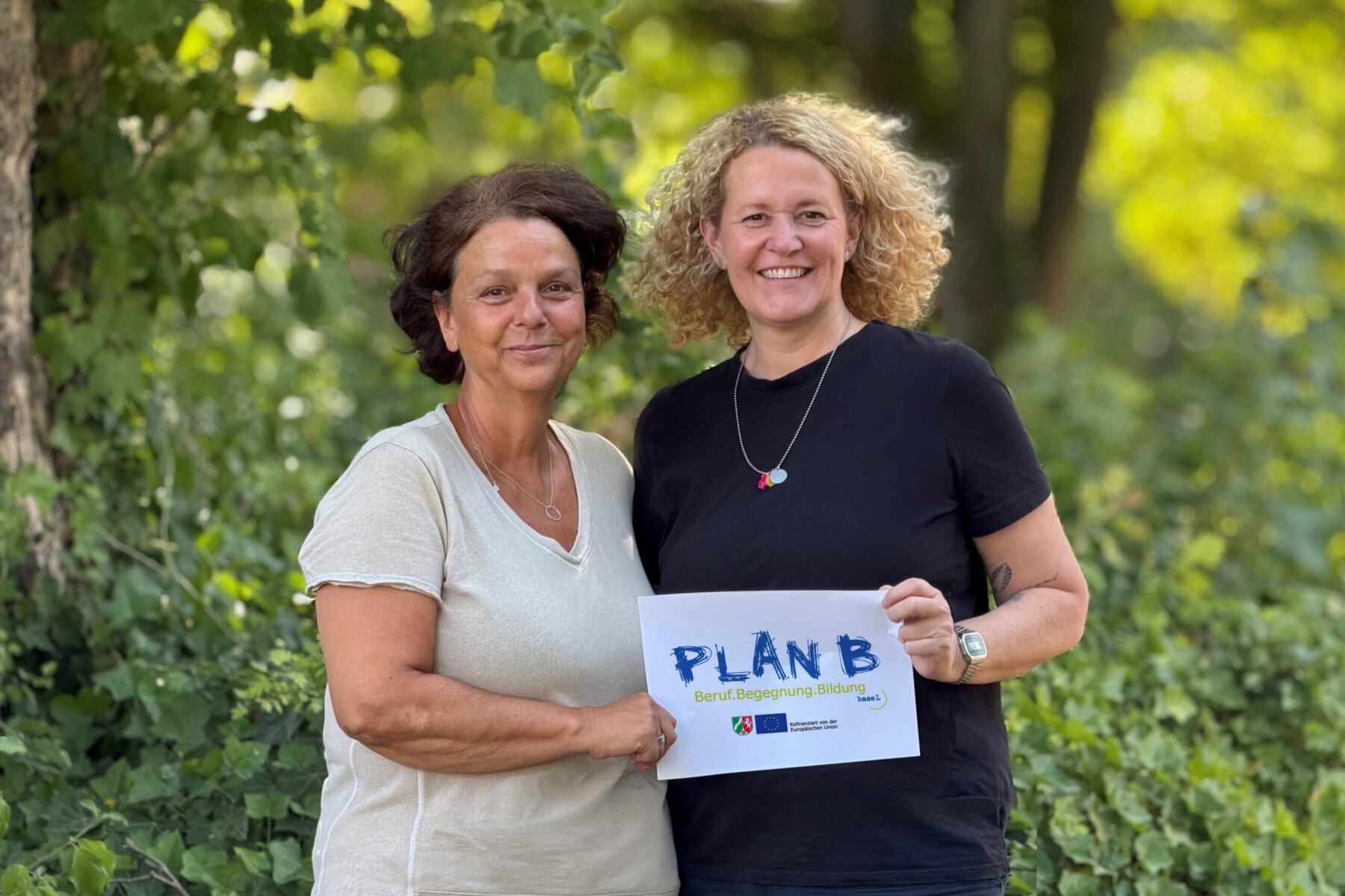 Zwei Frauen stehen vor Bäumen und halten ein Schild mit Text